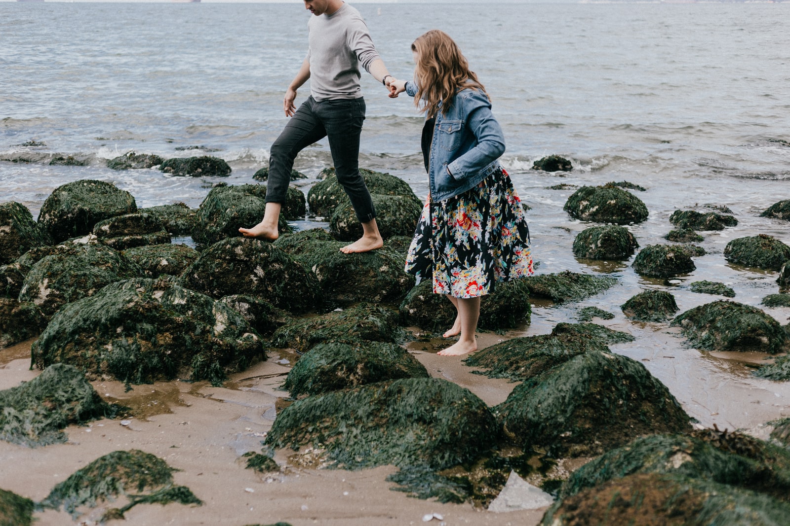 man and woman holding hand together while stepping on rocks near sea