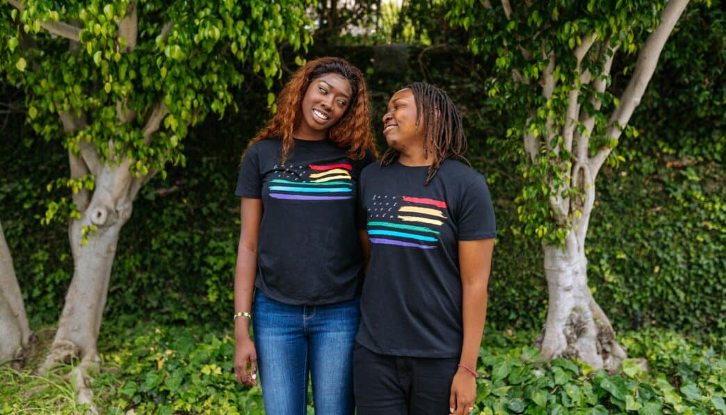 A joyful LGBTQ+ couple in matching Pride t-shirts, standing outdoors with trees in the background.