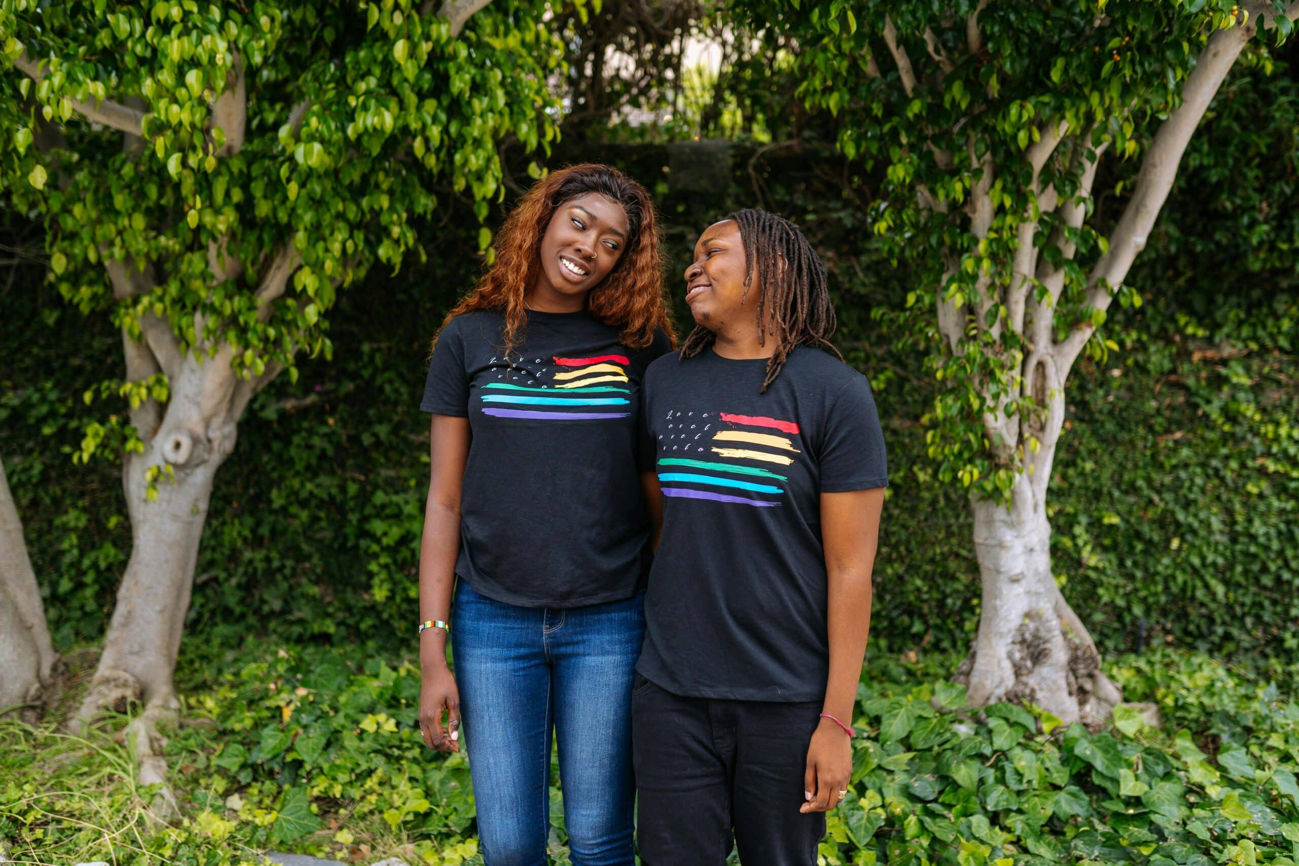 A joyful LGBTQ+ couple in matching Pride t-shirts, standing outdoors with trees in the background.