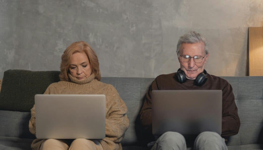 An elderly couple sitting on a sofa at home, each using a laptop. Modern technology engagement.