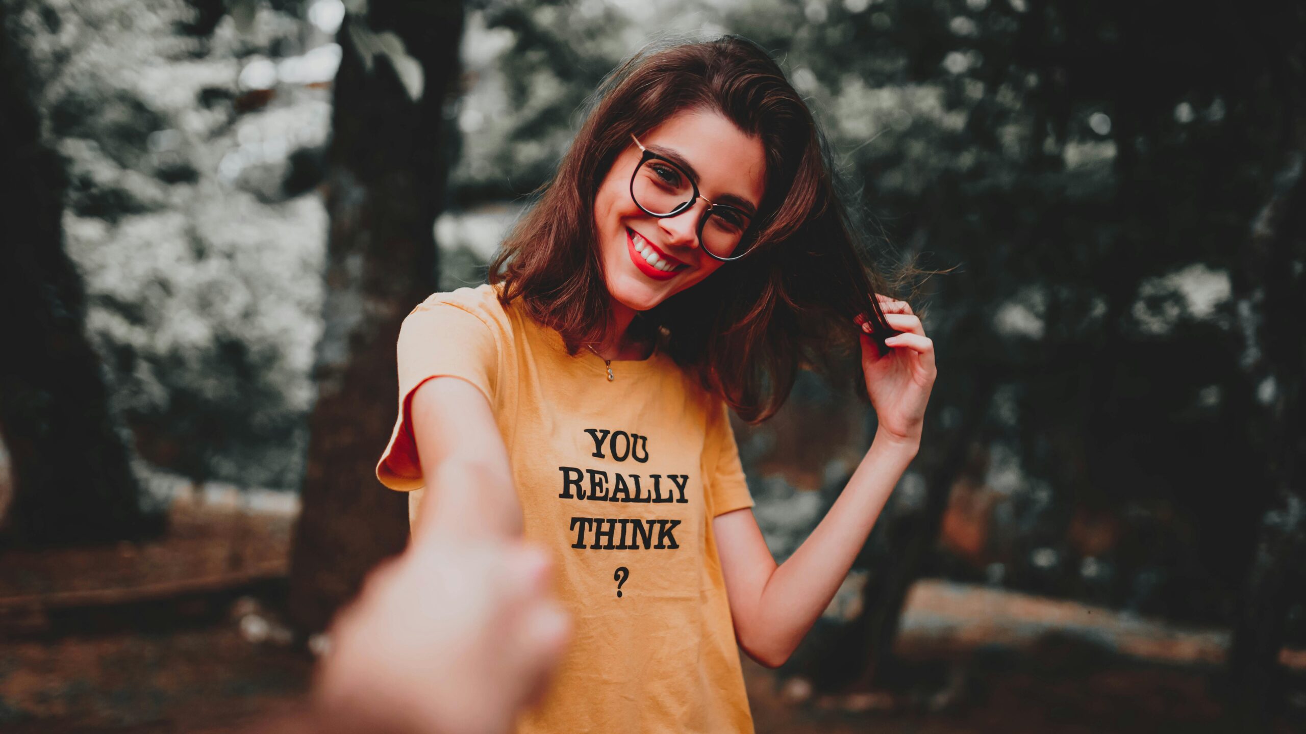 A cheerful woman in an orange shirt smiles playfully while holding her hair outdoors.