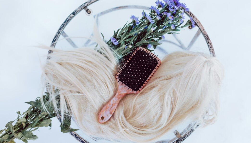 Blonde wig styled with brush and purple flowers on a round table, showcasing hair beauty and care.