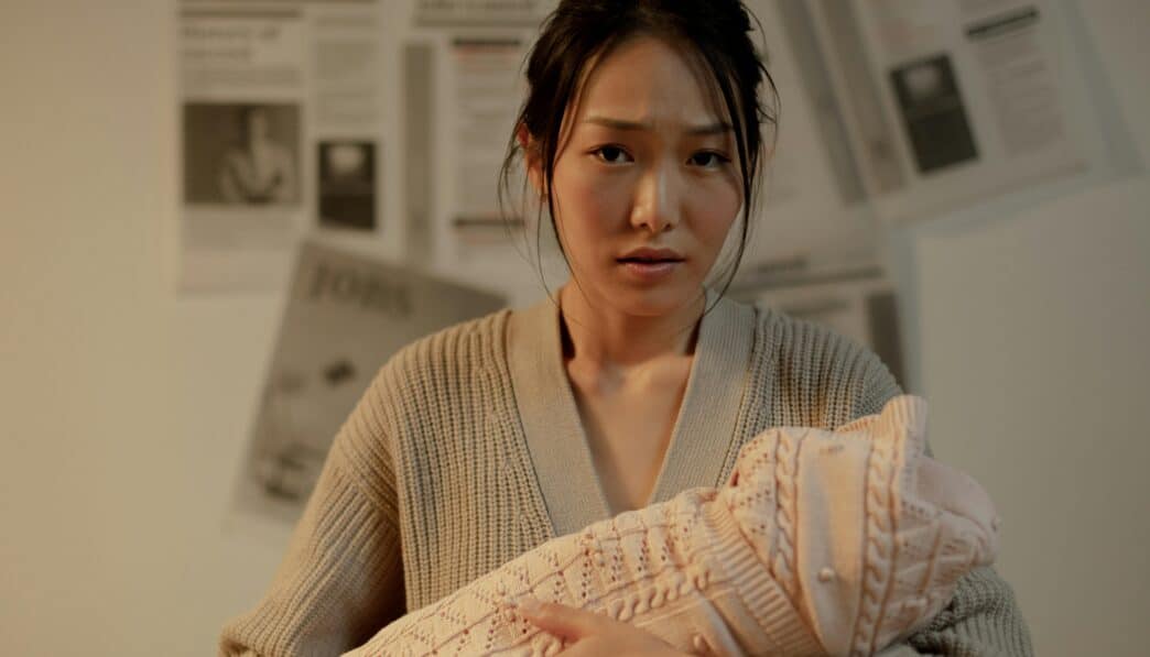 A concerned mother embraces her newborn indoors, surrounded by newspaper articles.